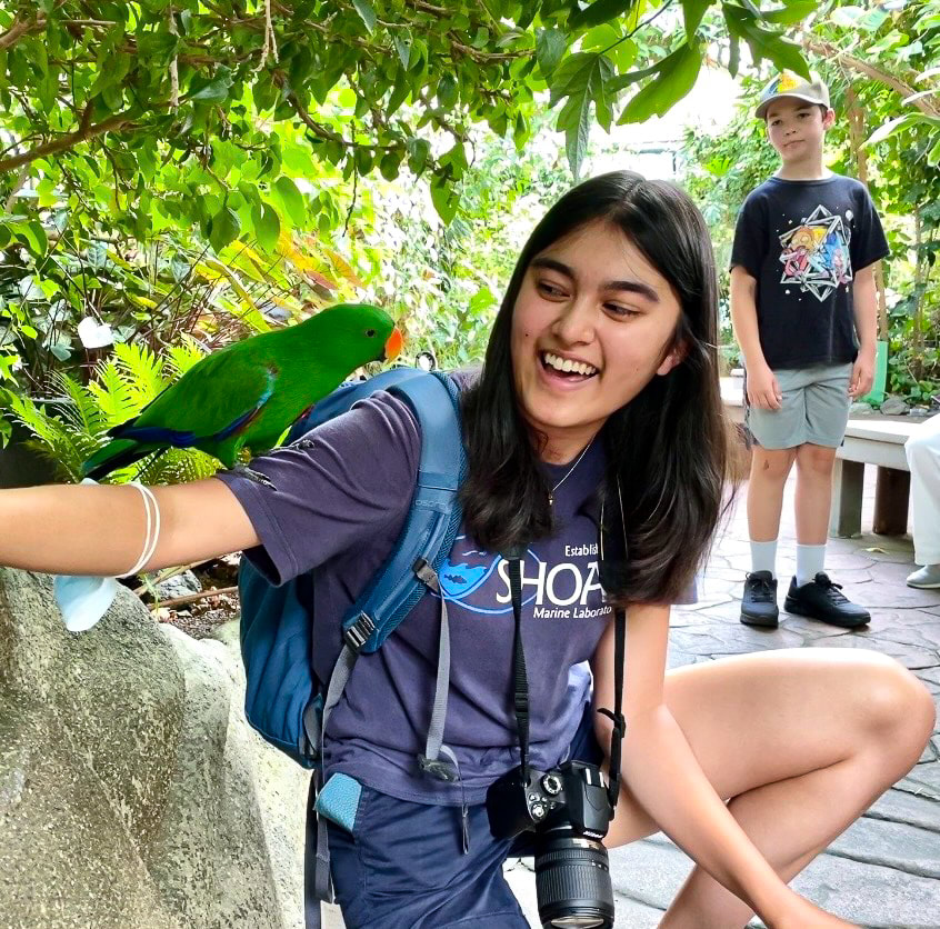A photo of Maya Xu with a camera around her neck and a parrot on her arm